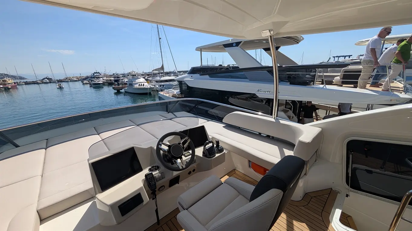 Interior view of a modern yacht cockpit featuring a steering wheel, navigation screens, and a scenic marina with moored boats.